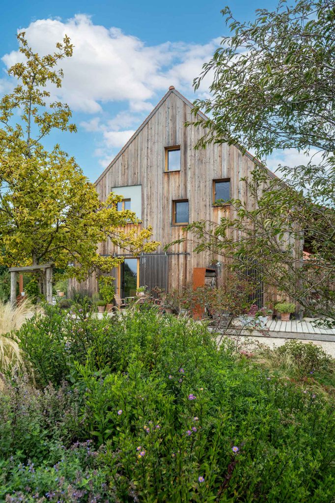 erneut das mit Holz beschichtete Haus, die Seite auf der man das Haus sieht zeigt viele unsymetrissche kleine Fenster und die Terasse