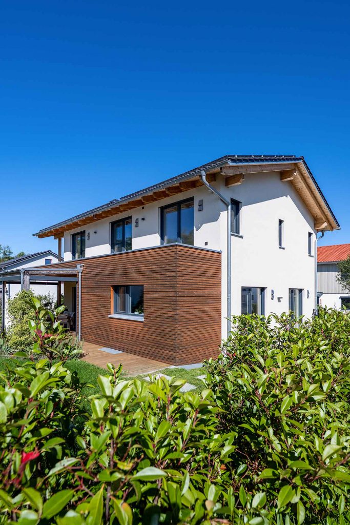 erneut die Rückseite des Hauses , man erkennt das Braune Holzgerüst und ein kleinen Teil der Terasse, im Vordergrund steht eher das Gebüsch der Hecke