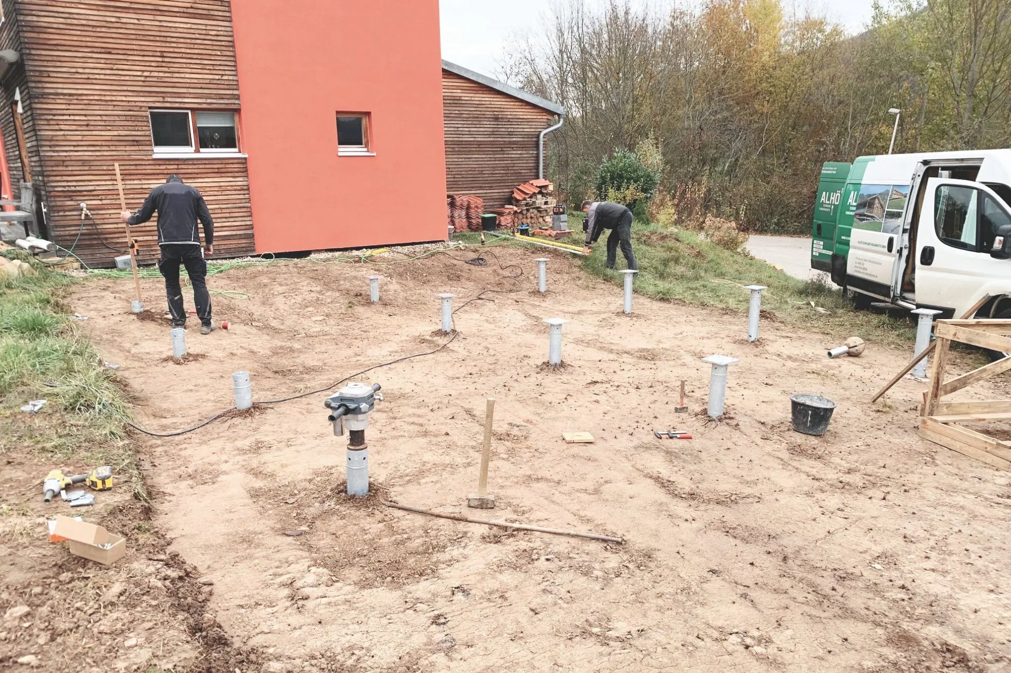 eine Baustelle mit Quadratischer Plattgemachten Erdfläche zu erkennen, man erkennt 2 in schwarz angezogene Bauarbeiter , einer von ihnen Hält eine Schaufel . Man erkennt auch Fundamentstützen in der Erdfläche