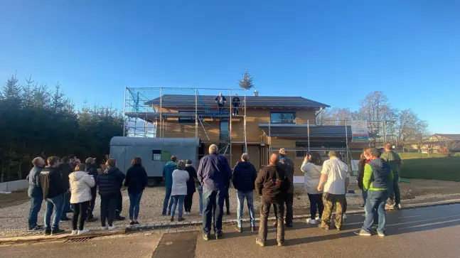 Eine große Menschenmenge vor einer Baustelle eines Familienhauses, auf der Baustelle 2 Männer auf dem Gerüst.