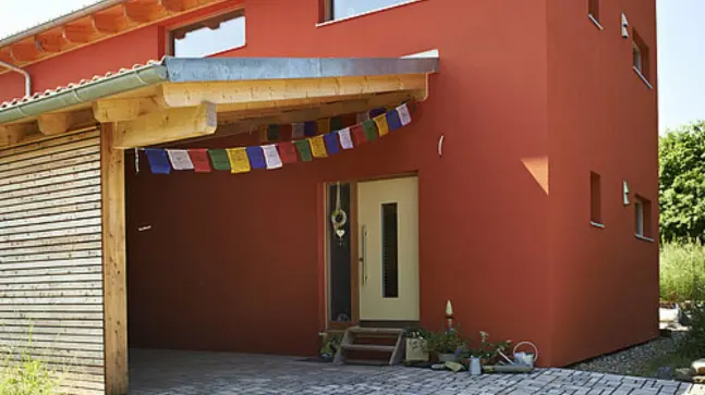 Rotes Haus mit einer Dachgaraga vor der Haustür was mit dem Haus verbunden ist, die Eingangstür ist Weiß und die Garage ist mit Bunten Flaggen beschmückt. Vor dem Haus erkennt man auch Dekoration wie Pflanzen und Gießkannen.