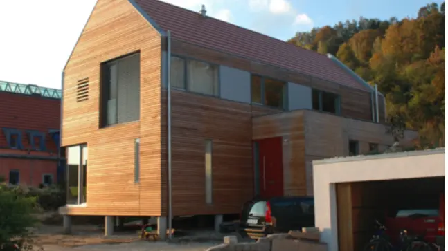 ein Großes Haus mit Rotem Dach. Das Haus ist von Außen mit Hellbraunen Holz beschichtet, es hat viele verschiedengroße und förmige Fenster und eine Rote Eingangstür. Das Haus liegt an einem Hochgelegenen Wald.