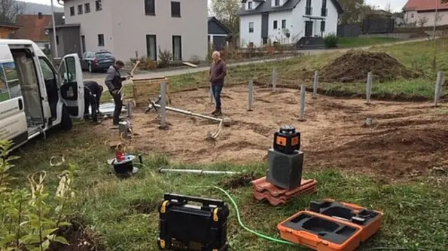 eine Baustelle, auf dieser erkennt man 8 Pfosten die in auf einer Quadtratischen Erdfläche fest sind. Auf dem Bild erkennt man auch 3 Bauarbeiter die ihren Beruf nachgehen. Ein Orangefarbiger Baukasten ist auch zu sehen