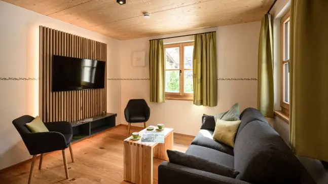 Art Wohnzimmer mit einer dunkelgrauen Coach mit grünen Kissen und einen Hellbraunen Holz Wohnzimmertisch, in dem Raum sind auch 2 Stühle und ein Fernsehr, hinter dem Fernseher ist eine Art Holzmuster und dahinter sind LEDS die Gelb leuchten