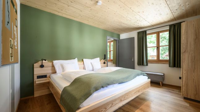 ein Grün Gestrichendes Schlafzimmer mit einem Doppelbett und zwei Nachtschränken, dieses Bett hat Weißen Bett überzug und eine Grüne Decke, das Schlafzimmer hat ein Fenster und einen Spiegel. Der Boden ist aus Dunklebraunen Holz