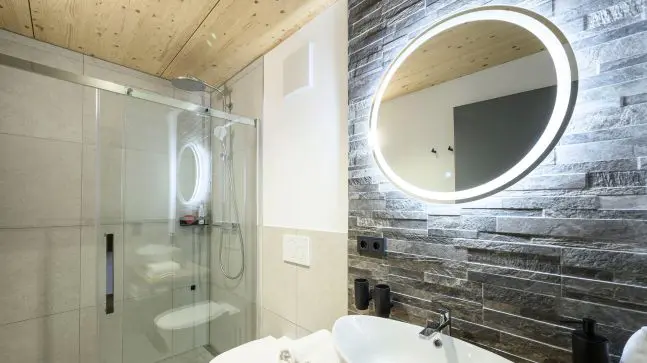 ein Bad mit Weisen Wänden und teilweise eine Steinoptik, man erkennt ein Runden Spiegel über dem Waschbecken , dieser hat einen LED Rahmen, eine Dusche ist in der rechten Ecke des Raumes zu erkennen.