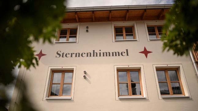 eine Hauswand mit der Aufschrift Sternenhimmel und 2 Roten Sternen daneben, das Haus besitz viele Fenster mit Holzgerüst