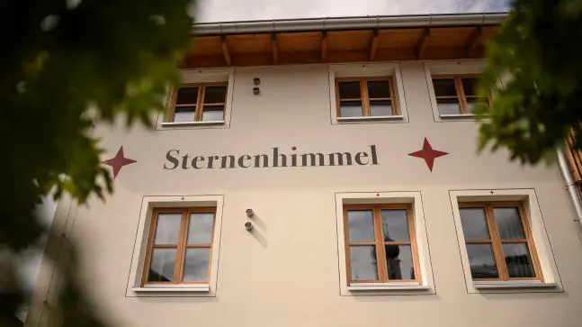 eine Hauswand mit der Aufschrift Sternenhimmel und 2 Roten Sternen daneben, das Haus besitz viele Fenster mit Holzgerüst