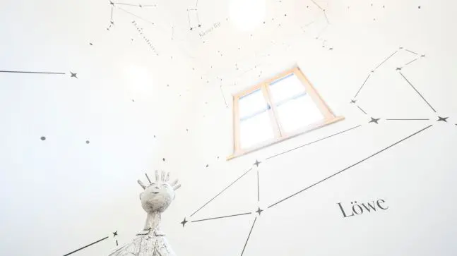 Raum mit Sternenhimmel und Sternzeichen aufgemalt, eine Weiße Kunstfigur und ein Braunes Holzfenster im Raum