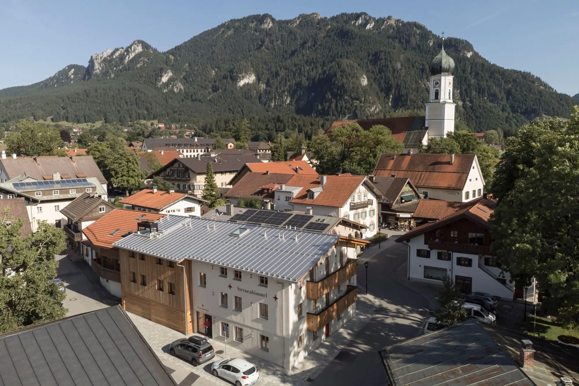 ein Dorf oder große Stadt zu sehen, sie liegt an einem Bewaldeten Berg und hat eine große Kirche mit Turkiser Spitze in der Stadtmitte, man erkennt viele Häuser mit roten oder blauen Dächern.